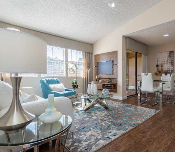 A living room with a white sofa, a blue armchair, a glass coffee table, and a television.