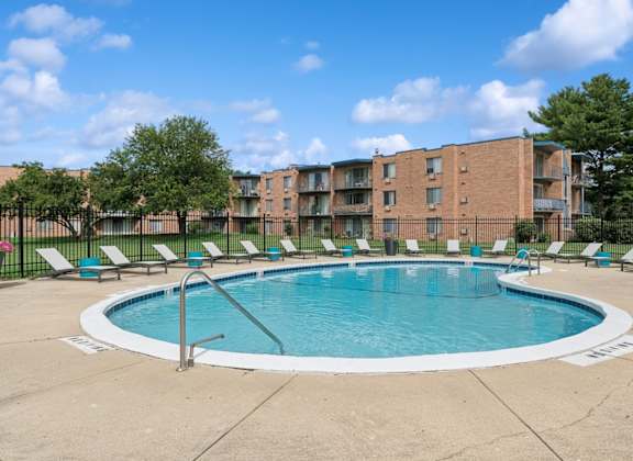 A swimming pool surrounded by lounge chairs and trees in front of a brick apartment building.