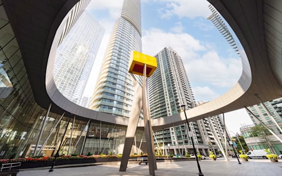 A large sculpture in the foreground with a yellow box on top stands in front of a modern building.