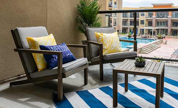 Two chairs with blue and yellow pillows on a balcony with a pool in the background.