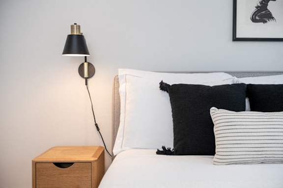 A lamp with a black shade is on a wooden stand next to a bed with white sheets and pillows.