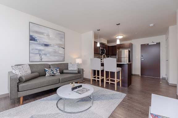 Living room area of an apartment, furnished with a sofa, rug, vinyl flooring, and high ceiling.