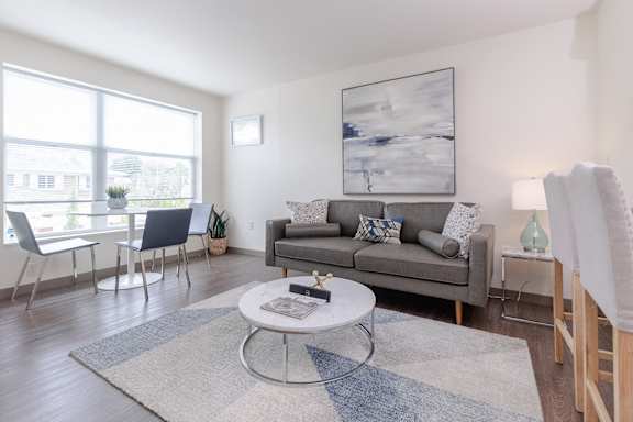 Living room area of an apartment, furnished with a sofa, rug, vinyl flooring, and high ceiling