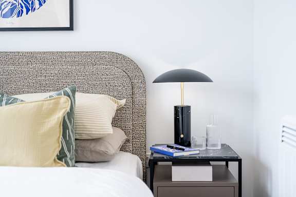 A lamp with a black shade sits on a small table next to a bed.