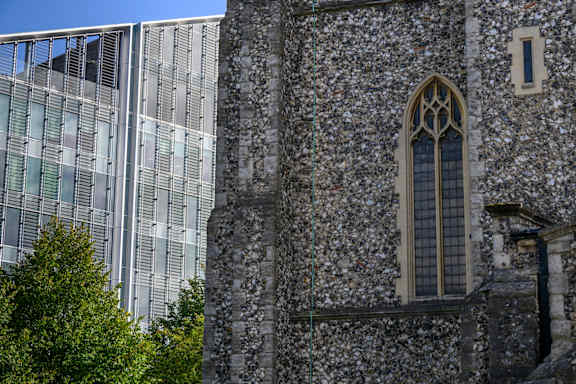 A tall building with a glass facade is behind a stone building with a large window.