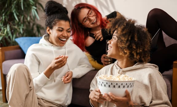 three women sitting on a couch eating popcorn and laughing