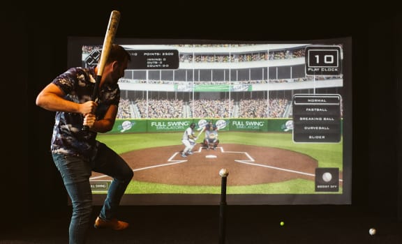 a man holding a baseball bat in front of a tv screen at The Apex at CityPlace, Overland Park, 66210