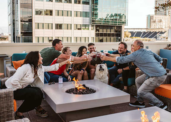 A group of people toasting with drinks around a fire pit on a rooftop.