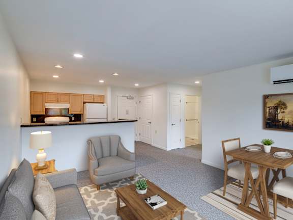 A living room with a grey couch, a wooden coffee table, a grey armchair, a wooden dining table with chairs, a painting on the wall, and a kitchen area in the background at The Birches at Chambers, New
