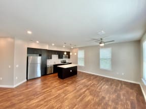 A large and open living space with 3 windows, hardwood-style flooring, and a ceiling fan leading a kitchen with stainless steel appliances.