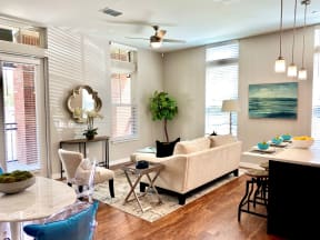 A living room with two windows, hardwood-style flooring, a ceiling fan, double doors leading to a private patio, and access to a dining area and kitchen.