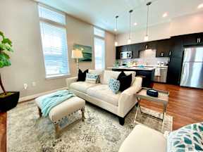 A living room with hardwood style flooring with two windows with open access to a kitchen with an island bar and hanging pendant lights.