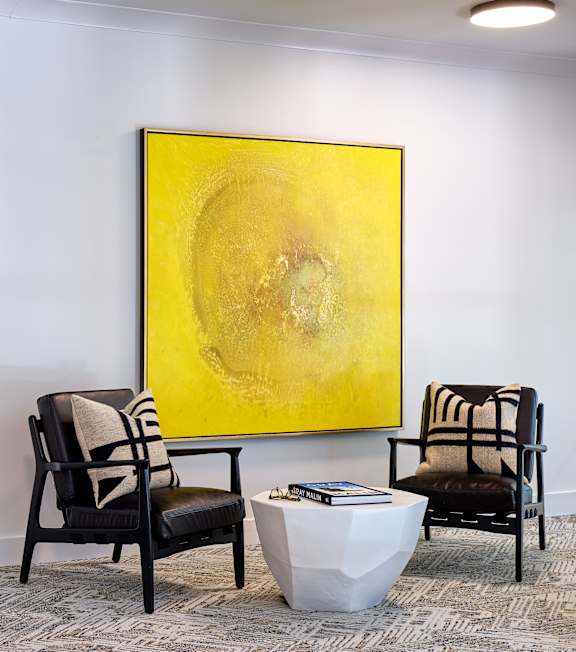 Two chairs and a table are in front of a yellow painting.