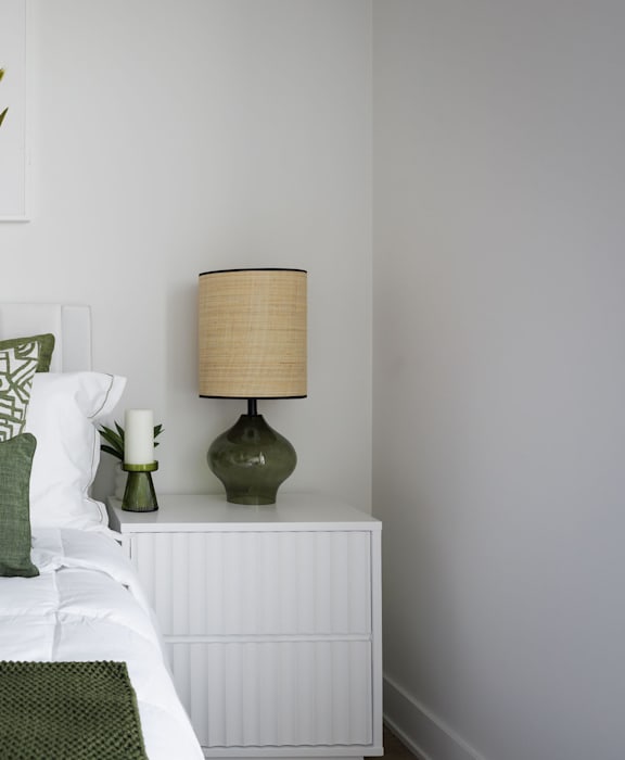 A white nightstand with a green lamp and a white bed with a green pillow.