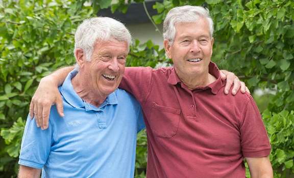 Two older men hugging each other in front of a green leafy background.