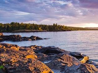 A serene landscape with a rocky shoreline and a forest in the distance under a twilight sky.