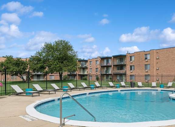 A swimming pool surrounded by lounge chairs and trees in front of a brick apartment building.