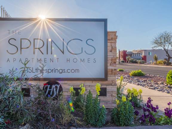 A sign for The Springs Apartment Homes is shown in front of a fence and some plants.