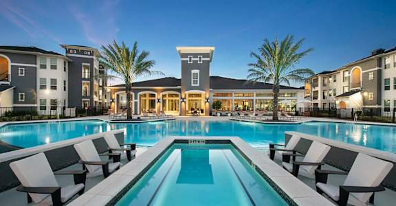 A large swimming pool surrounded by lounge chairs in front of apartment buildings.