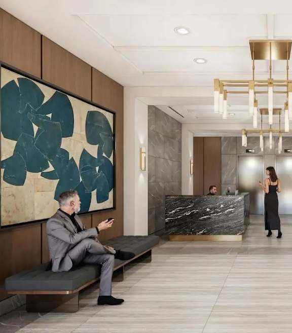 A man in a suit is sitting on a bench in a lobby with a woman standing in the background.