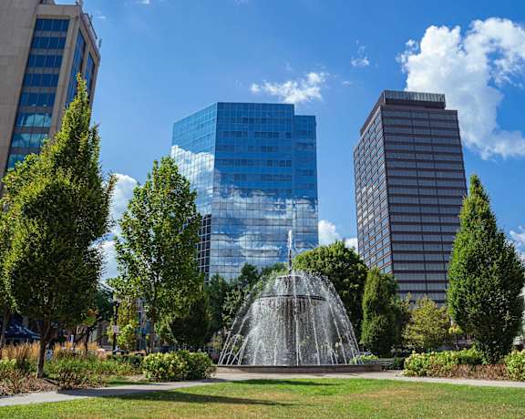 A fountain in the middle of a park with trees and buildings in the background.