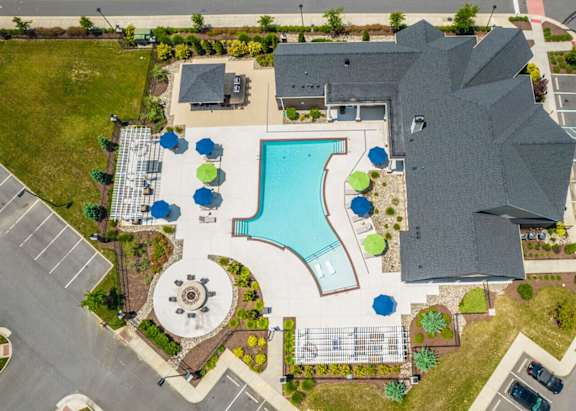 A large building with a grey roof is surrounded by a parking lot and a pool.