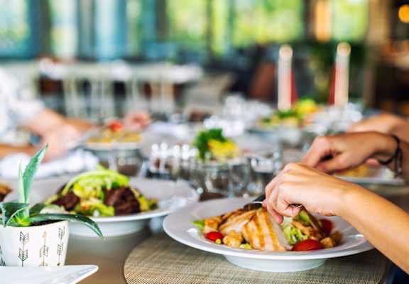 A person is cutting a sandwich on a plate at a table with other plates of food.
