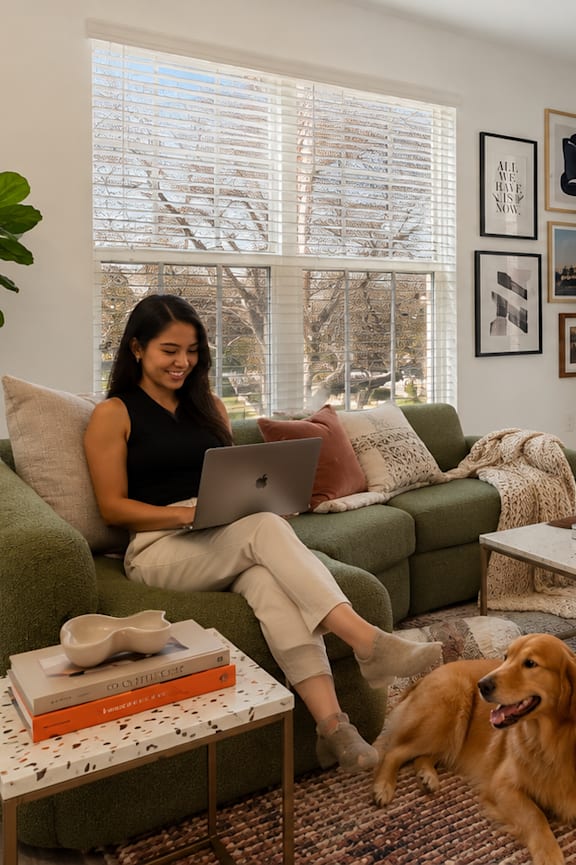 A woman is sitting on a couch with a laptop, a dog is standing next to her.