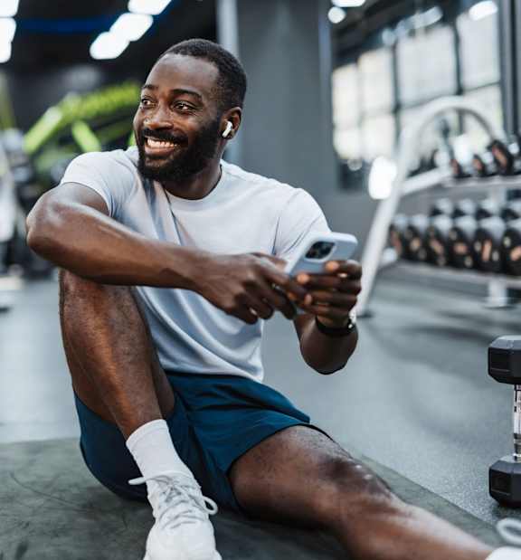 A man is sitting on the floor of a gym, looking at his phone.