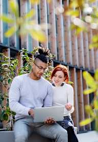 A man and woman are sitting on a bench with a laptop, surrounded by greenery.