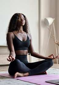 A woman is sitting cross-legged on a yoga mat in front of a laptop, meditating.