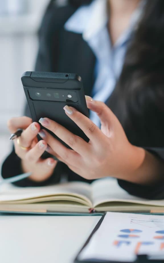 A person in a business suit is using a smartphone while sitting at a desk with a laptop and documents.