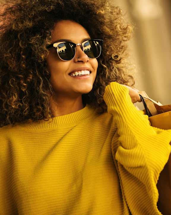 A woman with curly hair wearing sunglasses and a yellow sweater is holding shopping bags.