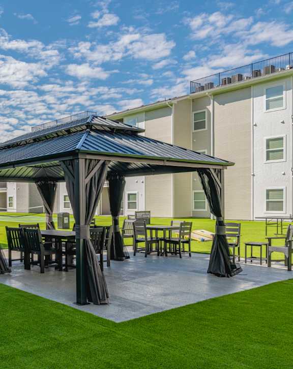 A gazebo with a table and chairs is set up on a grassy area in front of a building.