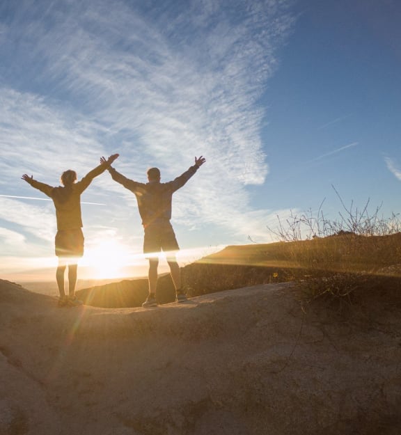 two people standing on a sand hill with their hands up in the air at sunset