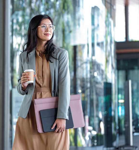 A woman in a brown dress and grey blazer is holding a cup and a pink folder.