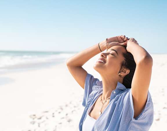 A woman in a blue shirt is standing on a beach with her hands behind her head.