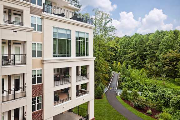 A modern apartment building with a balcony and a pathway leading to a wooded area.