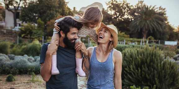 A man and woman are standing in a garden, with the man holding a child who is being lifted into the air by the woman.
