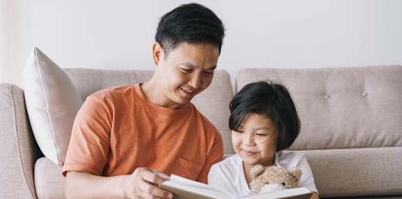 A man and a child are reading a book together on a couch.