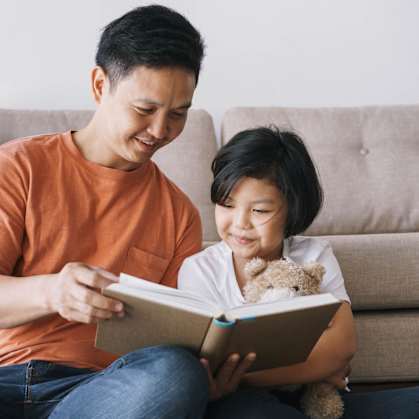 A man and a child are reading a book together on a couch.