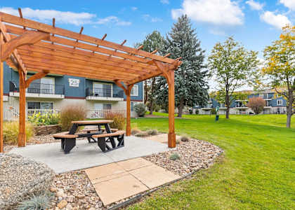 A wooden pergola with a picnic table is situated in a grassy area with a building in the background.