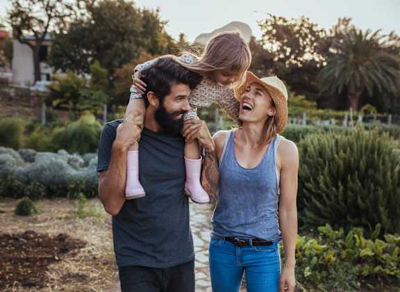 A man and woman are standing in a garden, with the man holding a child who is being lifted into the air by the woman.