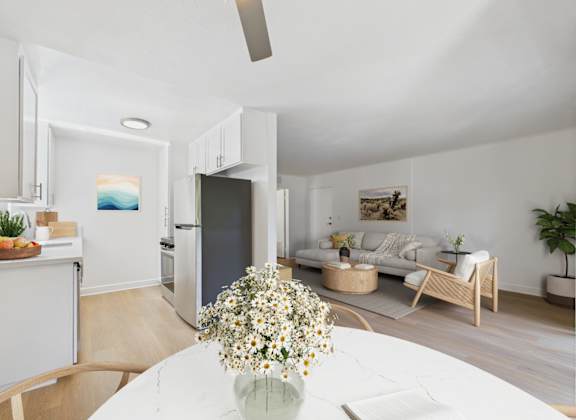 A kitchen with a refrigerator, a dining table with a vase of flowers on it, and a living room with a couch and a coffee table.