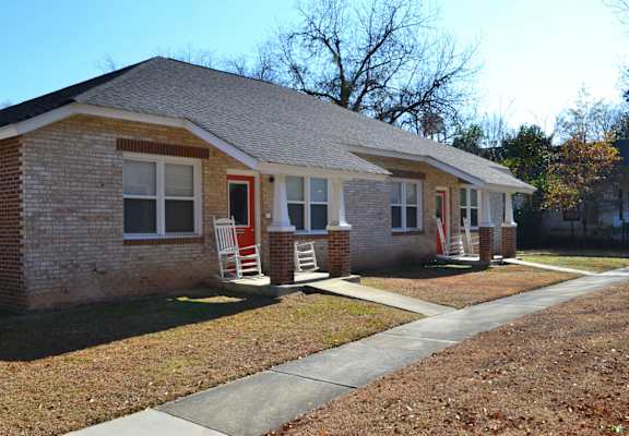 A small brick house with a red door and two white chairs in front.