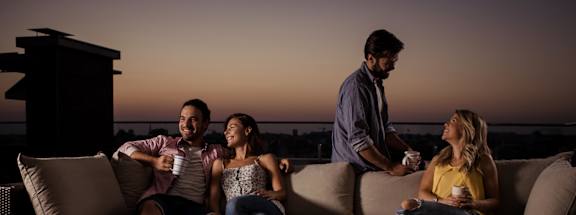 Four people sitting on a couch with a man standing and holding a tray of food.
