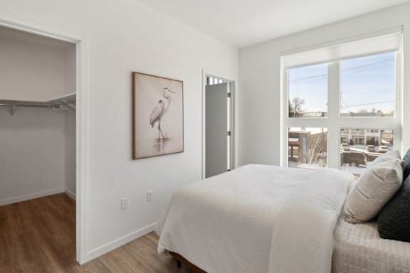A bedroom with a bed, a painting of a bird, and a view of the outside through a window.