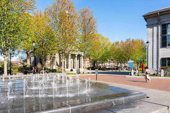 A fountain in the middle of a plaza with a woman walking in the background.
