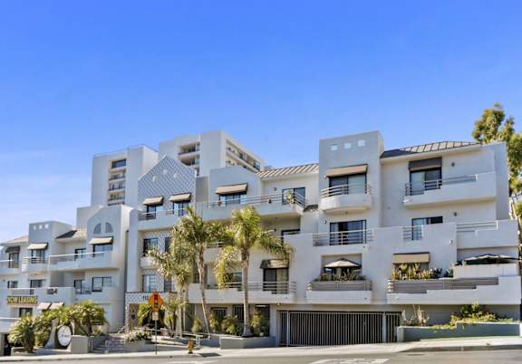 A modern white residential building with balconies and palm trees in front.