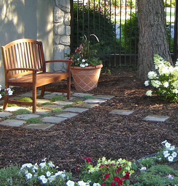 A wooden chair sits in a garden with a stone pathway leading to it.